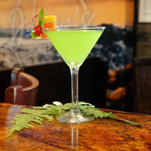 A vibrant green martini, garnished with a cherry and orange slice, sits on a glossy wooden bar.
