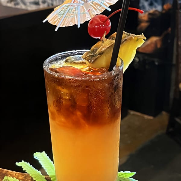 A tall glass of Mai Tai cocktail, garnished with a cherry, pineapple, and a paper umbrella.