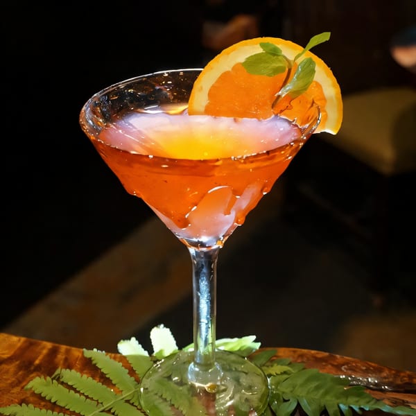 A vibrant orange cocktail in a martini glass, garnished with an orange slice and mint leaves.